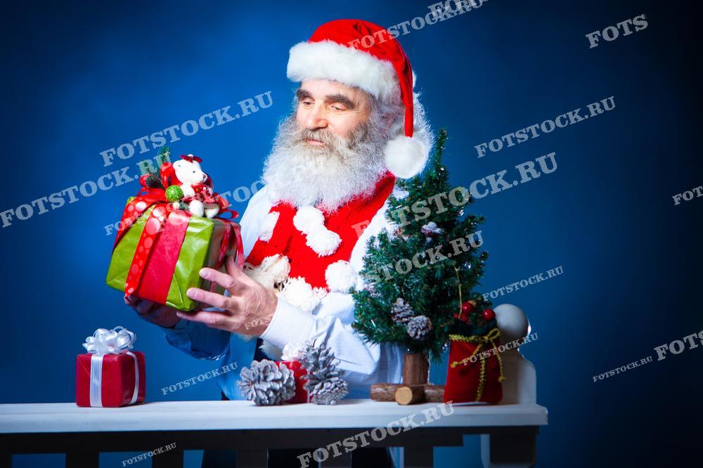 На изображении мужчина с седой бородой, одетый в шапку Санты и красный шарф, рассматривает праздничный подарок в яркой упаковке. Вокруг него рождественская елка и декорации, создающие праздничную атмосферу. Фотография символизирует рождественское настроение, ожидание праздника и обмен подарками.